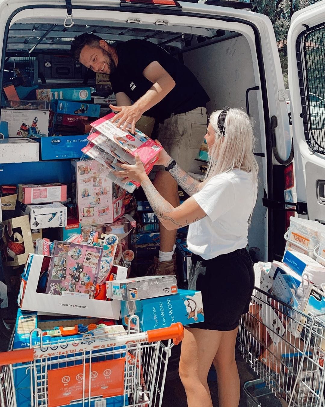 Two people unloading a van filled with boxes and items into a shopping cart.